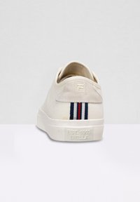 Sneaker bianca in tela con una texture liscia, caratterizzata da una patch del logo sul retro e tre strisce verticali in rosso, blu e nero sul tallone.