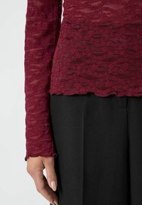 Top in pizzo bordeaux a maniche lunghe con motivi floreali texturizzati; bordi ondulati; indossato sopra un tessuto nero, parzialmente visibile sul fondo.