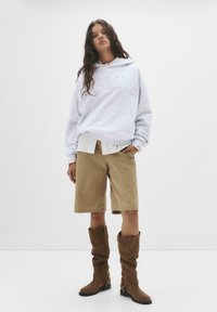 Felpa grigia con cappuccio e logo in rilievo, camicia bianca sotto, pantaloni corti beige e stivali slouch marroni alti. Sfondo semplice.