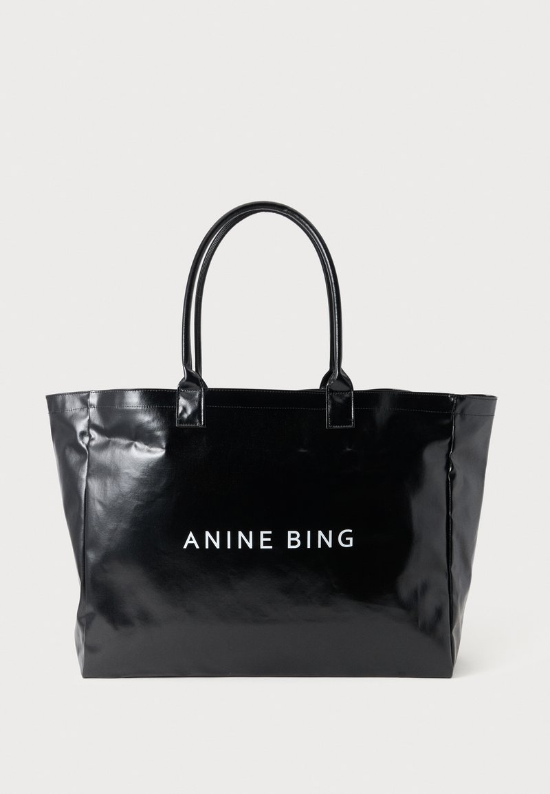 Sac fourre-tout noir brillant avec deux poignées et logo blanc "ANINE BING" centré à l'avant sur un fond clair uni.