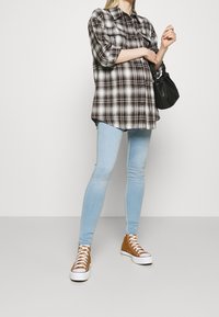 Camisa de cuadros en blanco y negro de gran tamaño con mangas enrolladas, jeans ajustados de color azul claro y zapatillas altas marrones, sosteniendo un bolso negro.