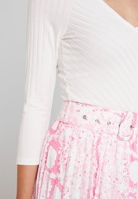 White ribbed top with a V-neck, paired with a pleated skirt featuring a pink and white abstract pattern and a matching belt with metal hardware.