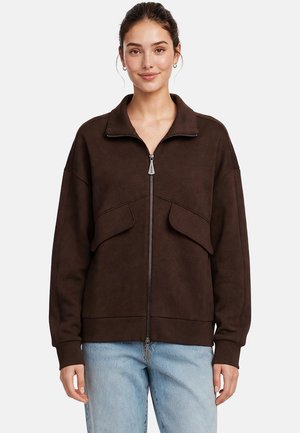 Donna che indossa una giacca marrone scuro con zip e tasche anteriori con patta e jeans azzurri chiari, in piedi davanti a uno sfondo semplice.