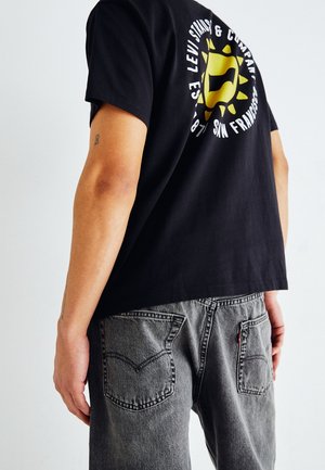 T-shirt en coton noir avec un logo soleil jaune et un texte blanc au dos, accompagné d'un jean en denim gris avec un design de poche classique.