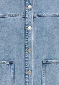 Tissu en denim bleu clair avec une rangée verticale de boutons-pression argentés et deux poches avant carrées avec coutures visibles.