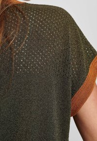 Strickoberteil mit einem strukturierten olivgrünen Oberteil und einem offenen Gewebemuster, ergänzt durch ärmel in kräftigem Orange.