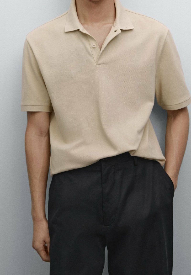 Homme portant un polo beige à manches courtes rentré dans un pantalon noir, debout devant un fond uni clair.