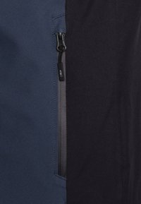 Marine en donkergrijs jacket sectie met een ritszak met een zwarte rits en een gemerkte trekker, zachte textuur en contrasterende kleuren.