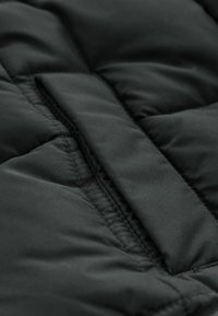 Jachetă puffer neagră dintr-un material cu textură matlasată. Prezintă o suprafață groasă și moale, cu cusături vizibile și un detaliu central cu clapetă.