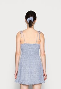 Vestido de estampado floral azul con tirantes ajustables, cuerpo fruncido y bajo con volantes. Un coletero a juego en el cabello añade detalle.