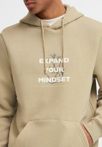 Sudadera beige con un gráfico frontal que presenta el texto "EXPANDE TU MENTE" en blanco, y elementos florales impresos arriba. Textura suave, bolsillo canguro.