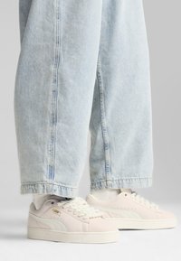 Tenisky Puma suede v svetloružovej farbe s bielymi akcentmi, s plochými šnúrkami a textúrovanou gumovou podrážkou, spárované s svetlomodrými širokými džínsami.