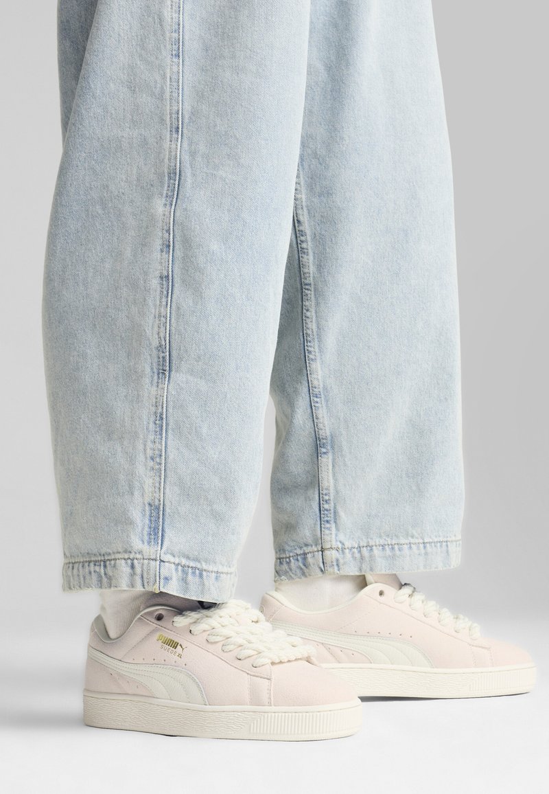 Tenisky Puma suede v svetloružovej farbe s bielymi akcentmi, s plochými šnúrkami a textúrovanou gumovou podrážkou, spárované s svetlomodrými širokými džínsami.