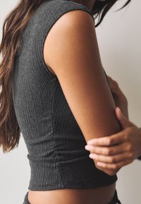 Haut sans manches gris à côtes avec un design ajusté, présentant un tissu texturé et un détail froncé à la taille. Le bras du mannequin repose sur le côté.