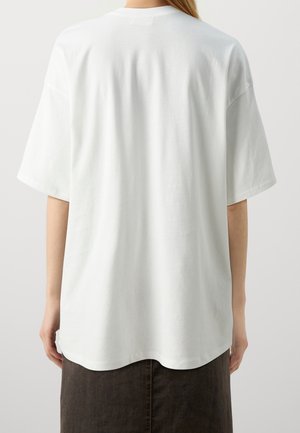 Person, der står med ryggen til iført en simpel hvid oversize kortærmet t-shirt og en mørk nederdel mod en ensfarvet baggrund.