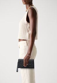 Black leather clutch with studded design, featuring a zipper and wrist strap, held in a hand. Light-colored, ribbed knit top and pants visible.