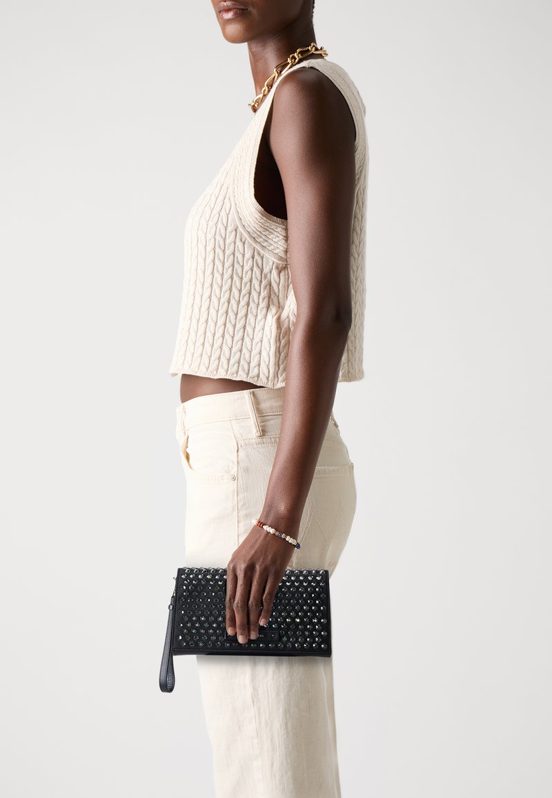 Black leather clutch with studded design, featuring a zipper and wrist strap, held in a hand. Light-colored, ribbed knit top and pants visible.