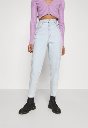 Osoba ubrana w jasnoniebieskie jeansy z wysokim stanem, czarne sznurowane buty i fioletowy prążkowany top z długim rękawem i krótkim krojem na jednolitym tle.