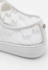Talons de baskets blancs Michael Kors avec tige en mesh transparent présentant des logos MK répétés et nom de marque embossé sur la semelle.
