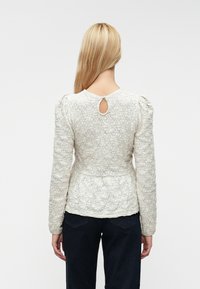 Blusa texturizada em marfim com mangas longas, cintura ajustada e detalhe em forma de fenda nas costas. O tecido apresenta um padrão floral. Combinada com jeans escuros.