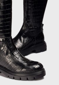 Botas negras hasta la rodilla con un exterior de cuero con grabado de cocodrilo texturizado, suela de goma robusta y fondo grueso con relieve para mayor tracción.