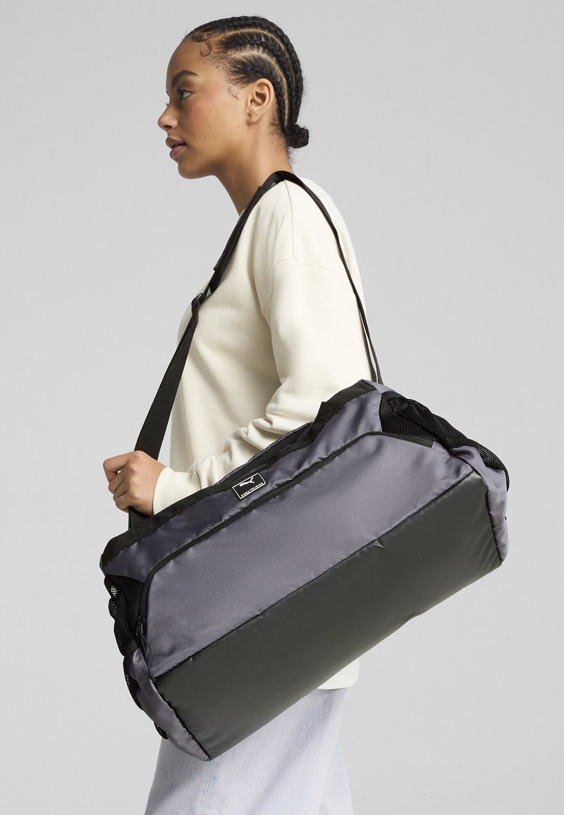 Sac de sport noir et gris en tissu lisse, doté d'une bandoulière, d'une fermeture éclair et d'un accent de logo. Poches latérales en maille texturée.