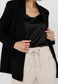 Schwarzer Blazer mit taillierter Passform, kombiniert mit einem schwarzen Satin-Camisole und beigen Stoffhosen mit Zugband und seitlichen Fransen.