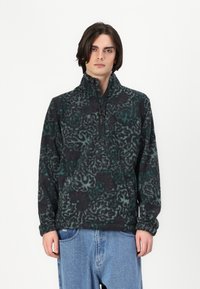 Fleece-pullover i mørkegrå med grønt abstrakt bladmønster, halv-glidelås krage og lange ermer. Myk tekstur og avslappet passform.