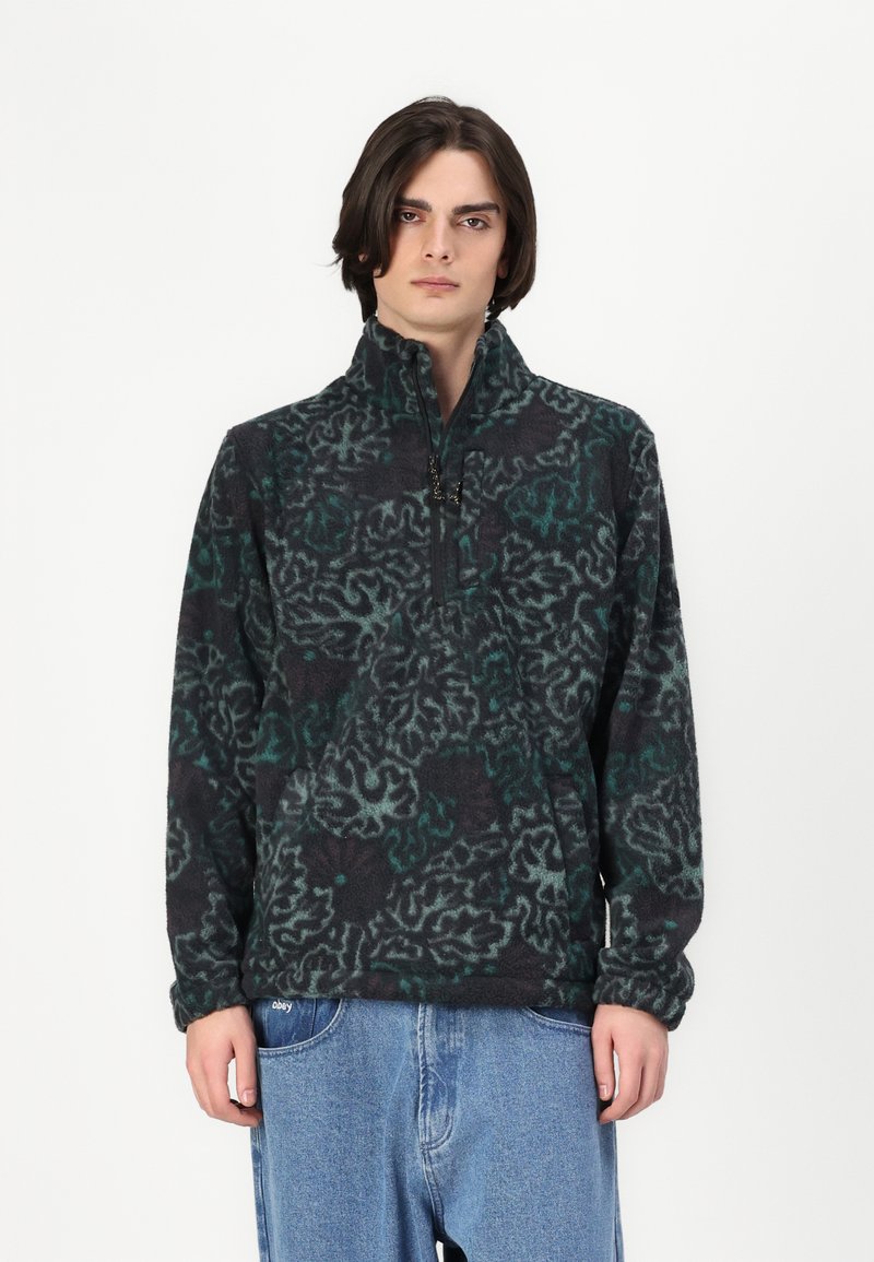Fleece-pullover i mørkegrå med grønt abstrakt bladmønster, halv-glidelås krage og lange ermer. Myk tekstur og avslappet passform.