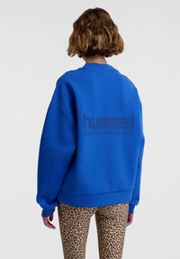 Blaue Sweatshirt mit oversize Schnitt, Rundhalsausschnitt und dunkler Schrift "hummel PERFORMANCE" auf dem Rücken. Getragen mit Leggings im Leopardenmuster.
