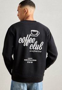 Schwarzer Pullover mit weißem Drucktext auf der Rückseite: "Coffee Club frisch und heiß serviert" mit einer Tassen-Grafik und Branding darunter.