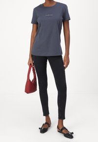 Tmavě šedé bavlněné tričko s krátkými rukávy a logem na hrudi, v kombinaci s černými úzkými džínami a černými balerínami, držící červenou kabelku.