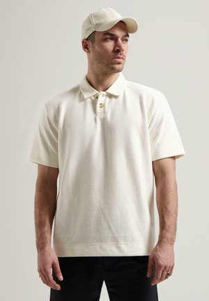 Polo shirt bianco a trama con maniche corte e colletto a doppio bottone. Abbinato a un cappello bianco coordinato. Il modello è in piedi davanti a uno sfondo neutro.