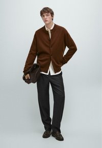 Brauner Wollcardigan mit rundem Ausschnitt und schwarzen Knöpfen, über einem weißen Hemd getragen. Graue maßgeschneiderte Hose und dunkle Schuhe, dabei eine braune Tasche haltend.