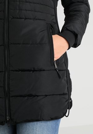 Gros plan sur une personne portant une veste matelassée noire, une main glissée dans une poche zippée et un jean bleu visible.