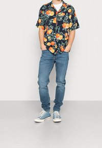 Camicetta nera a maniche corte con motivi floreali e fruttati arancioni e gialli, abbinata a jeans in denim blu e sneakers azzurre chiare.