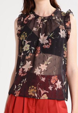 Blouse noire transparente sans manches à imprimé floral avec épaules volantées, portée sur un top noir, associée à un pantalon rouge taille haute.