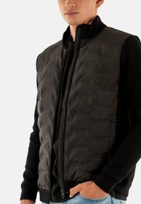 Gilet nero con cerniera, texture imbottita, collo alto e tasche laterali, indossato sopra una maglia nera a maniche lunghe. Design semplice, materiale leggero.