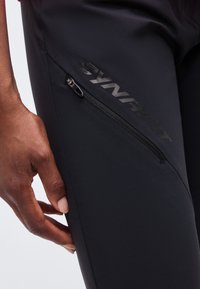 Κοντινό ενός χεριού που ακουμπά κοντά στην τσέπη ενός μαύρου παντελονιού με διαγώνιο φερμουάρ και λογότυπο DYNafit στο μηρό.