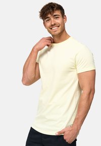 Camiseta de algodón amarillo claro con mangas cortas, cuello redondo y corte ajustado, que presenta un sutil logo en el pecho. El modelo posa con la mano en el cuello.