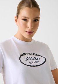 Weißes T-Shirt mit schwarzem, besticktem Logo in einem ovalen Seildesign, auf dem "GUESS EST 1981" steht, mit Rundhalsausschnitt und kurzen Ärmeln.