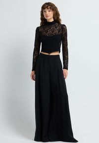 Mustapitsi crop-top, jossa on pitkät hihat, korkea kaulus ja monimutkaiset kuviot, yhdistettynä leveälahkeisiin mustiin housuihin.