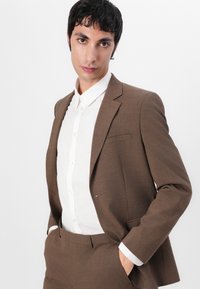 Sacou din stofă maro, ajustat, cu un singur buzunar, purtat peste o cămașă albă cu nasturi. Costumul are un material texturat și croială dreaptă.