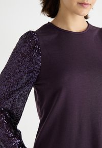 Top violet avec un corps lisse et une manche bouffante en sequins de couleur violet foncé, mettant en valeur la texture et le scintillement du design de la manche.