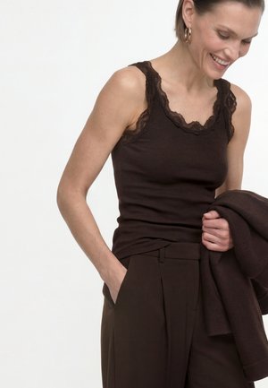 Mujer sonriendo y mirando hacia abajo, lleva una camiseta sin mangas marrón oscuro con encaje y pantalones a juego, sosteniendo una chaqueta marrón oscuro.