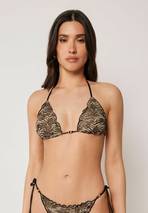 Kvinde iført gennemsigtig zebra-print mesh bikini med bindesnore og gyldne perleaccents, stående mod en ensfarvet lys baggrund.