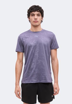 Młody mężczyzna stoi, ubrany w fioletowy t-shirt w wzory i czarne spodenki z logo Puma na jasnym tle.