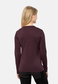 Donna con lunghi capelli biondi, che indossa una maglietta a maniche lunghe di un viola scuro e pantaloni neri, vista di spalle su uno sfondo chiaro e semplice.