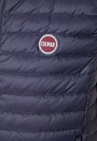 Chaleco acolchado azul marino con patrón de costura horizontal, con un logo circular de Colmar en burdeos y blanco en el área del pecho izquierdo.