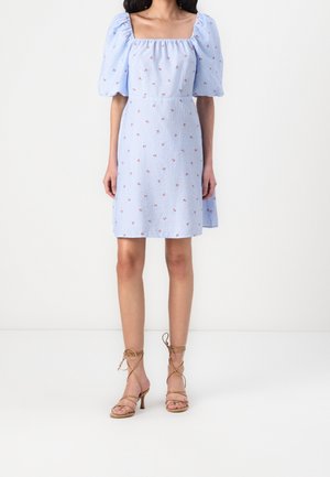 Mujer con un vestido azul claro con mangas abullonadas y pequeños estampados florales rojos, combinado con sandalias de tacón alto en tono camel con tiras.